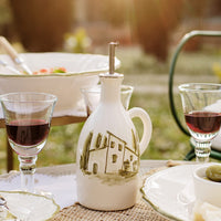 Ceramic olive oil dispenser with house design on a table with glasses of red wine and plates outdoors.